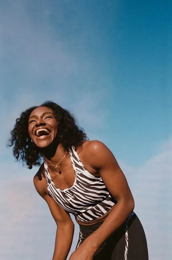 Mulher rindo, inclinada para a frente depois do treino, com o céu azul ao fundo