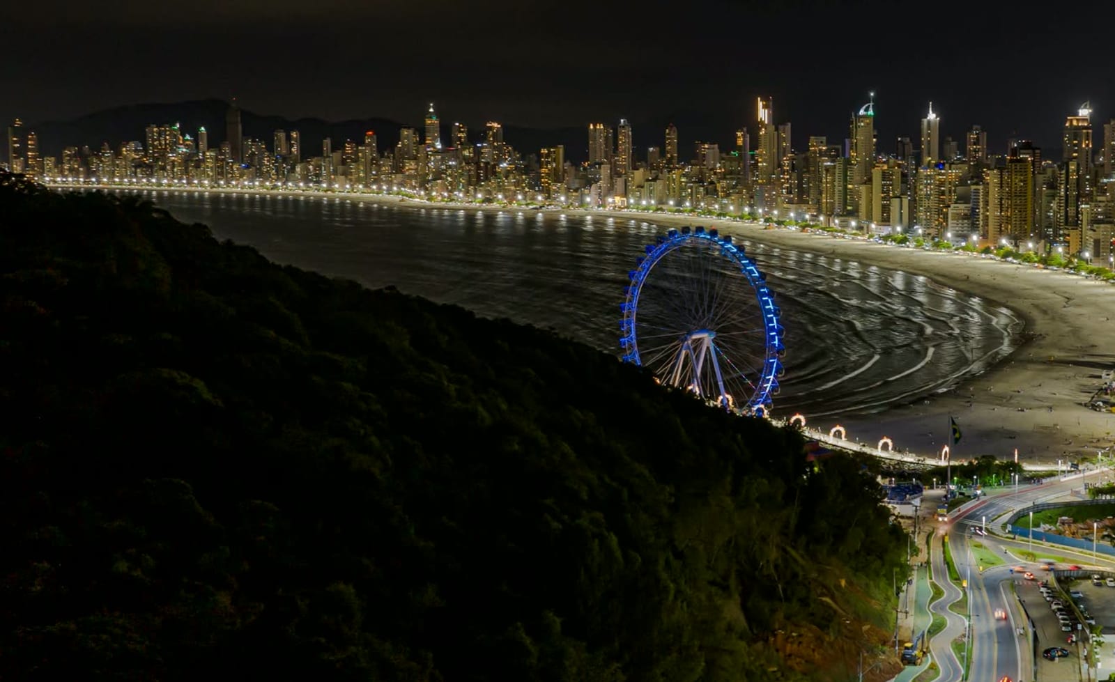 Orla de Balneário Camboriú à noite, o skyline iluminado ao fundo da baía e a roda-gigante azul junto à praia