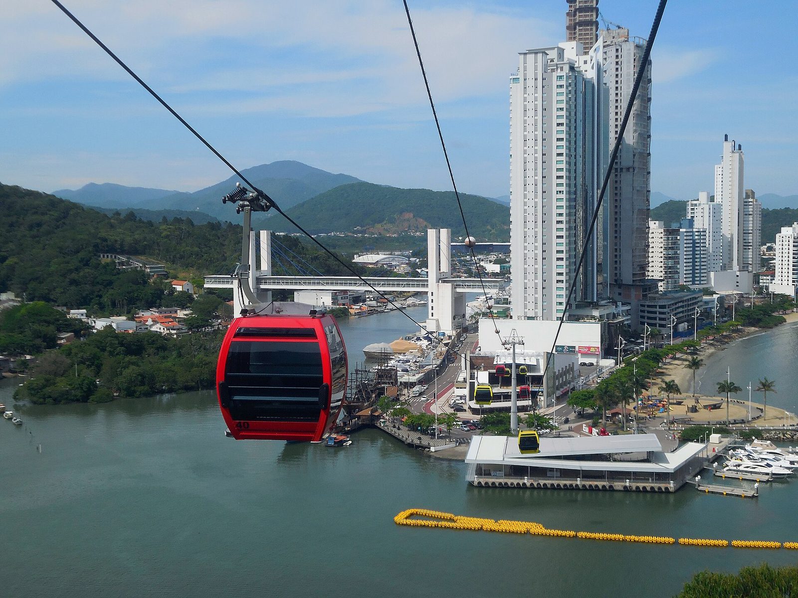 Bondinho vermelho do teleférico do Parque Unipraias cruzando o canal de Balneário Camboriú de dia, com a estação Barra Sul e os prédios da orla ao fundo