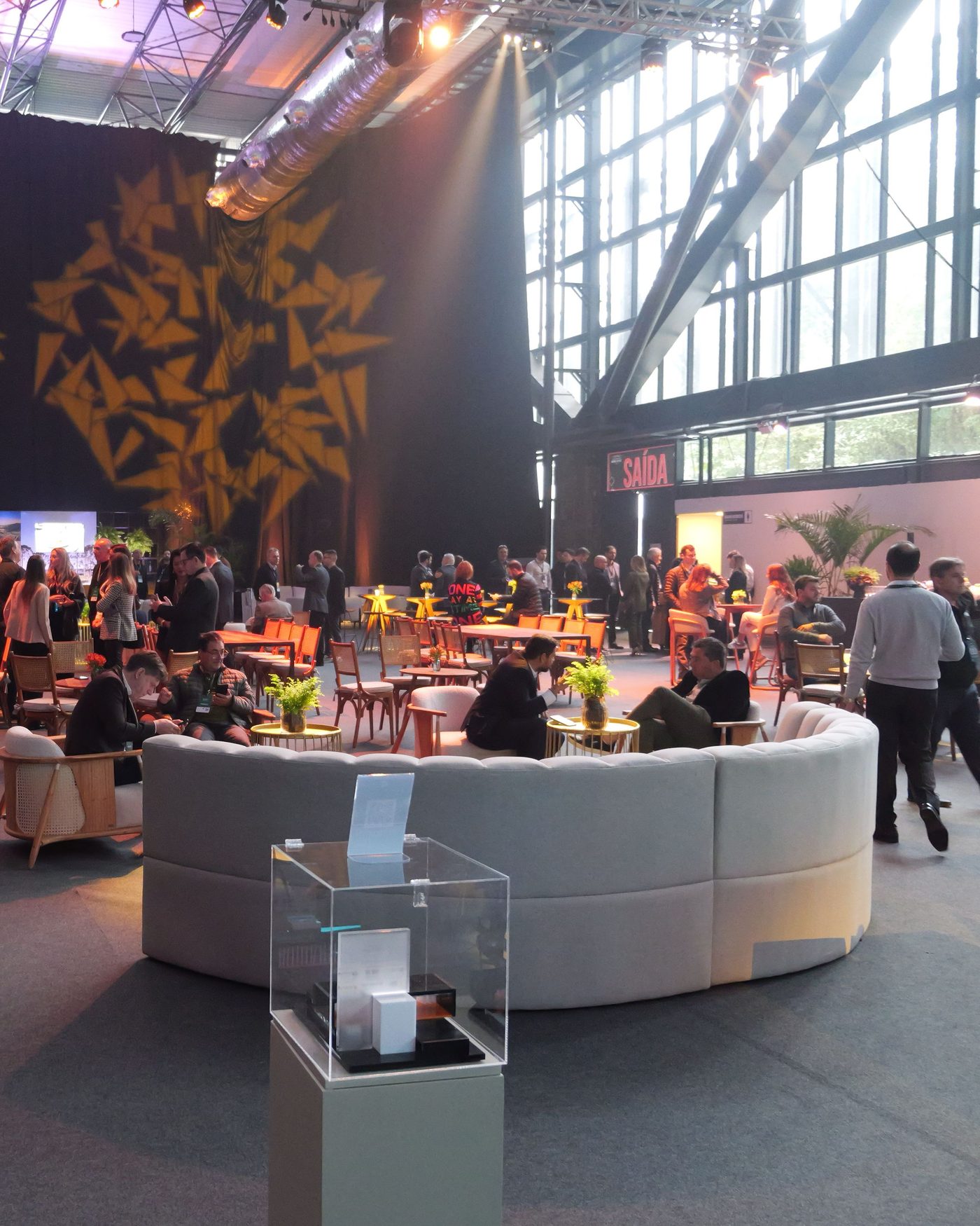 Área de convivência montada dentro do pavilhão do Expocentro, com sofás, mesas e público circulando sob a estrutura metálica do telhado