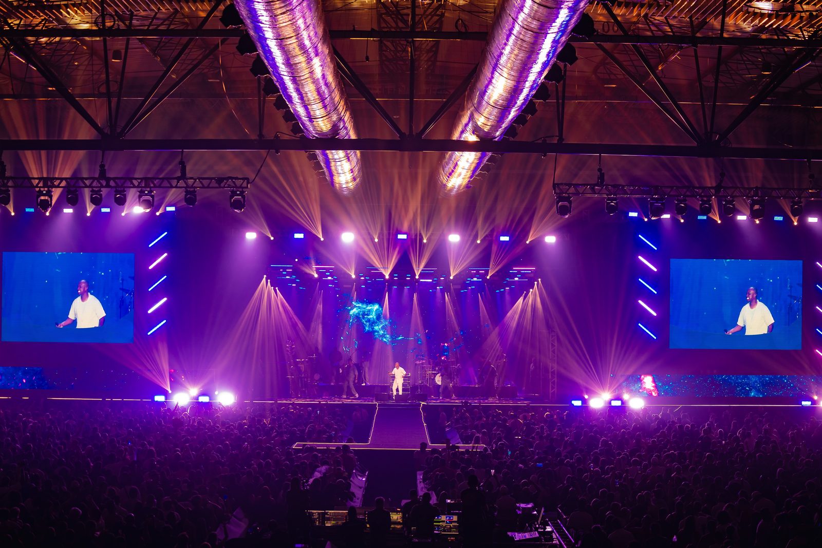 Show com o palco iluminado em luzes roxas e douradas, plateia acompanhando de pé em um pavilhão de eventos