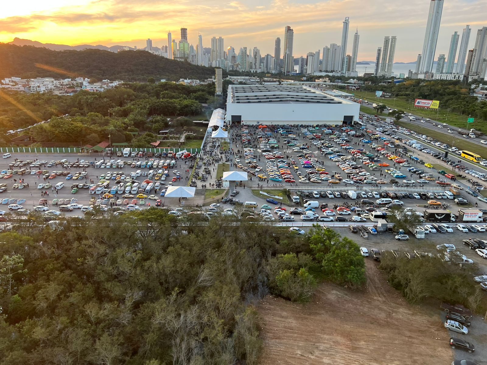 Vista aérea do Expocentro no fim da tarde, com o pátio tomado de carros e público e a orla de Balneário Camboriú ao fundo