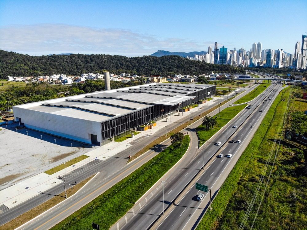 Vista aérea do Expocentro Balneário Camboriú, o pavilhão à beira da rodovia com o skyline da cidade ao fundo