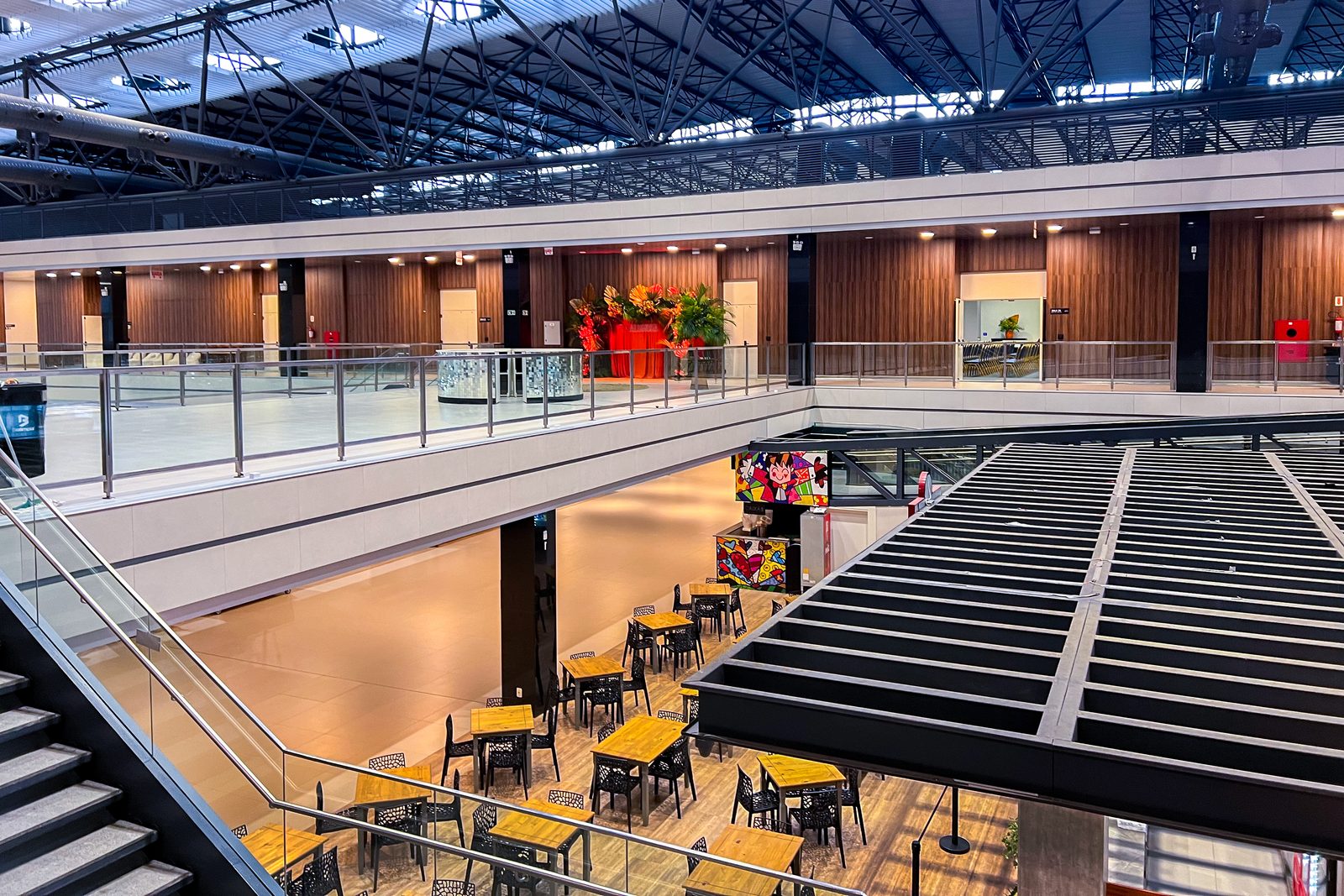 Interior do Expocentro Balneário Camboriú: mezanino em volta do vão central, treliça metálica no pé-direito e a praça do térreo com mesas de madeira