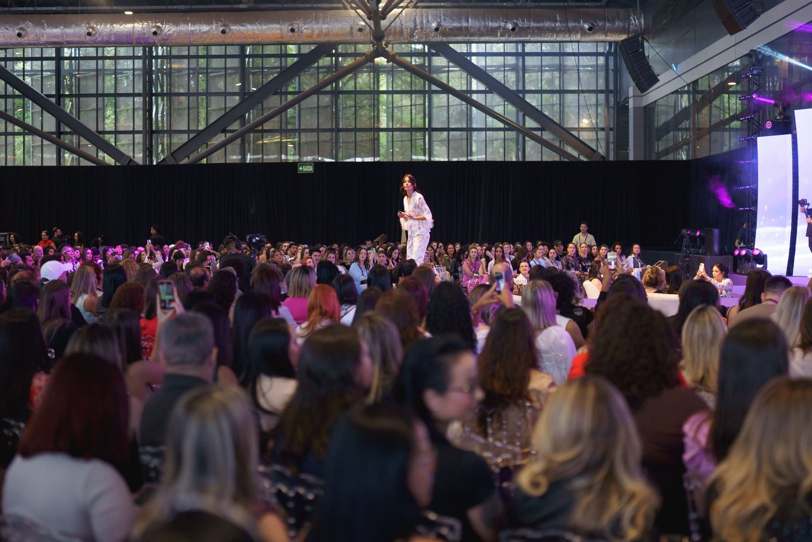 Pavilhão do Expocentro lotado durante um evento, com o público sentado diante da passarela e a fachada de vidro ao fundo