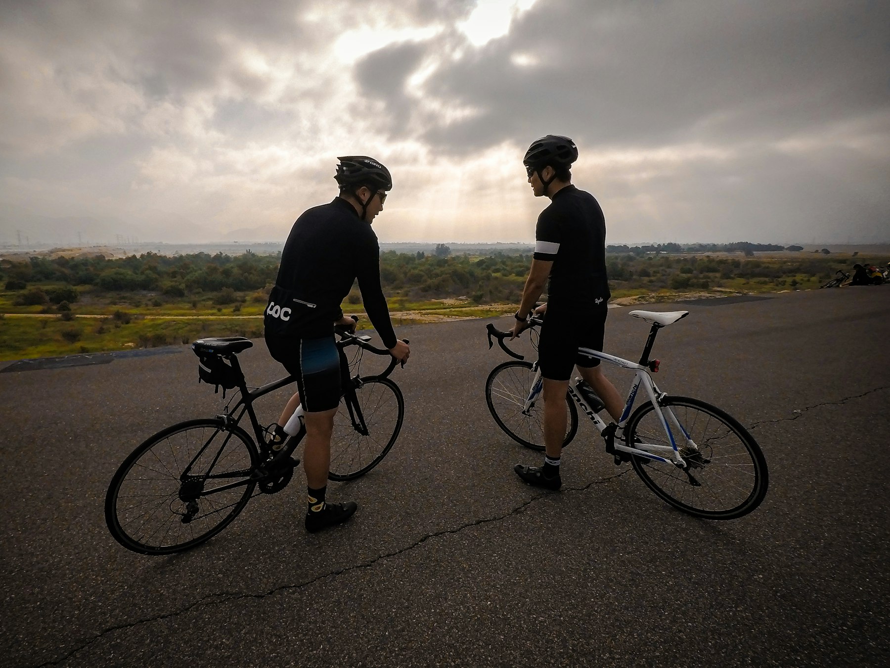 Dois ciclistas de estrada conversam parados no asfalto, com o sol atrás das nuvens