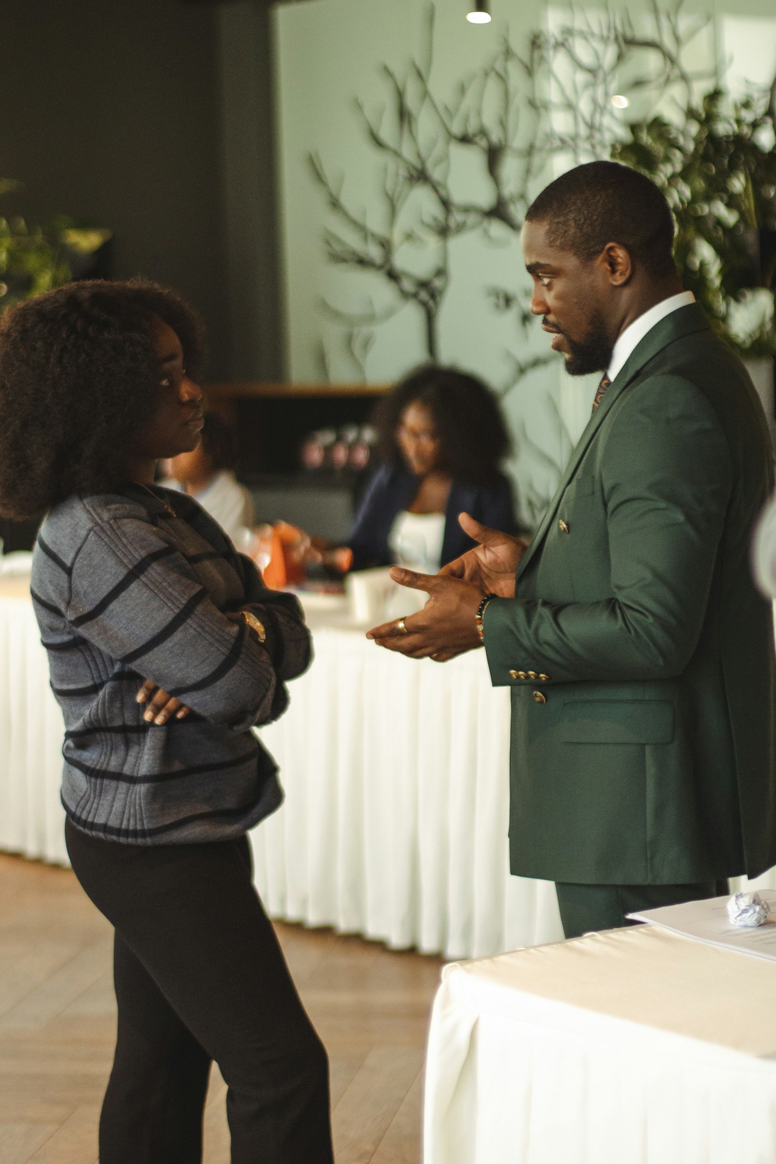 Duas pessoas em trajes de trabalho conversando em pé durante um evento, uma delas explicando com as mãos