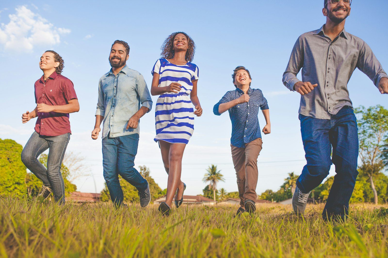 Grupo diverso de amigos correndo e sorrindo juntos num campo de bairro, com telhados e uma palmeira ao fundo
