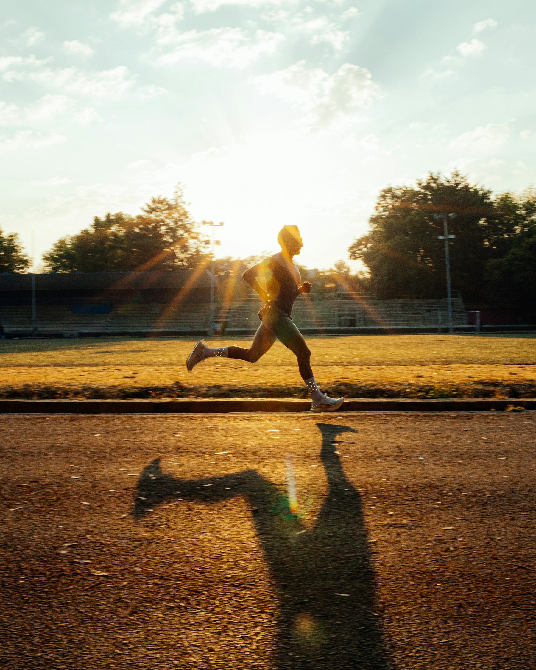 Homem correndo em uma pista de atletismo ao pôr do sol, silhueta contra a luz, arquibancada vazia ao fundo