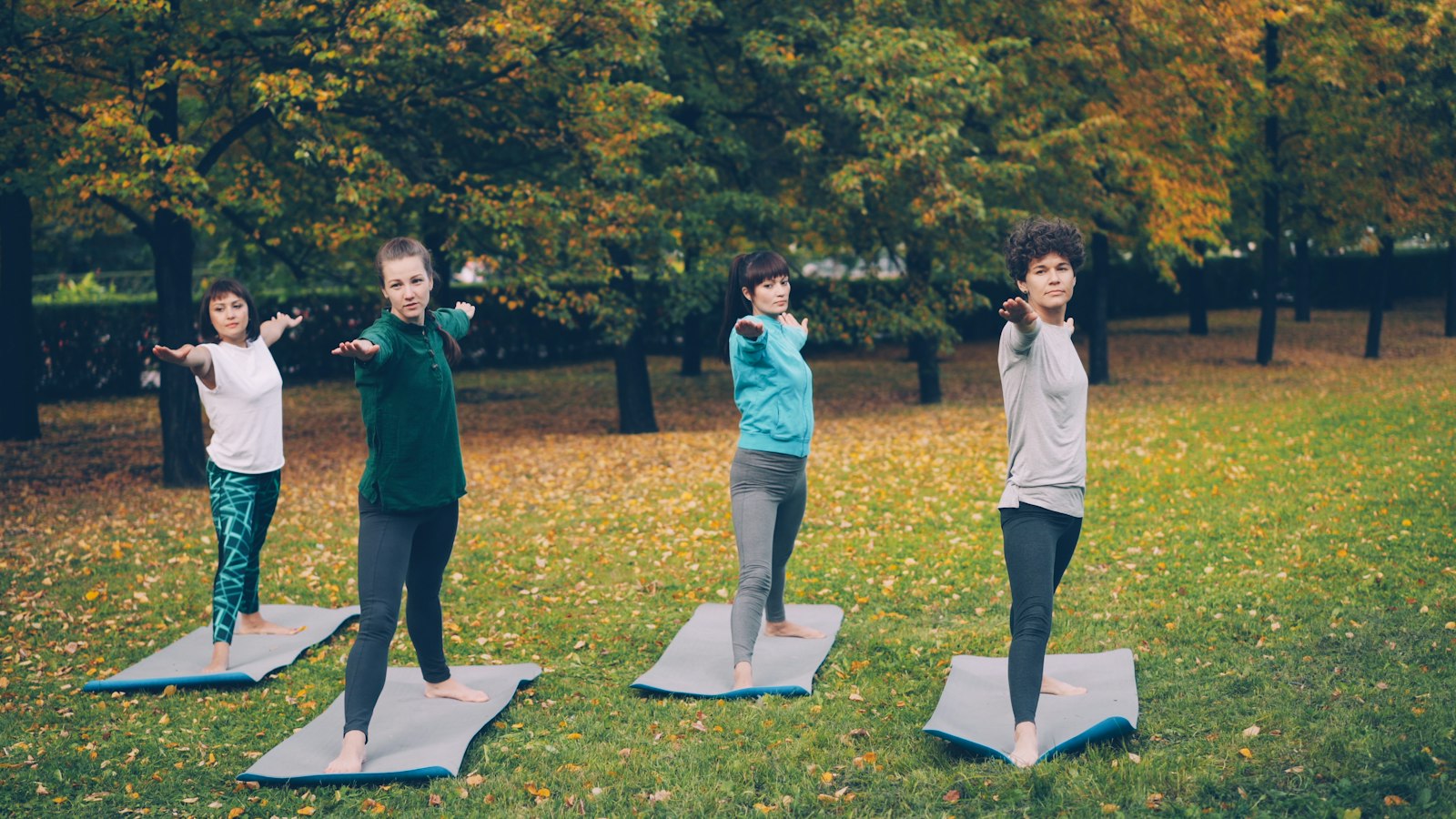 Quatro mulheres num parque, em pé sobre tapetes, praticando juntas um movimento de alongamento com o braço estendido à frente