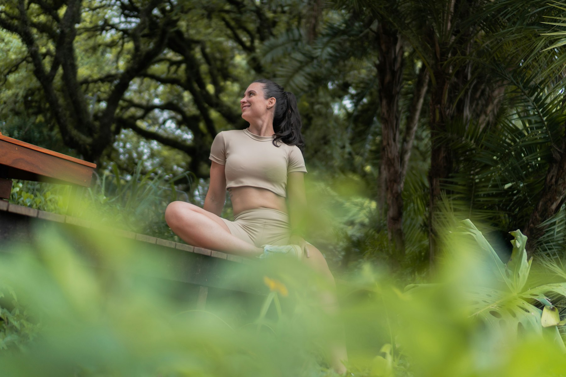 Mulher sentada de pernas cruzadas ao ar livre, cercada de vegetação, olhando para o alto