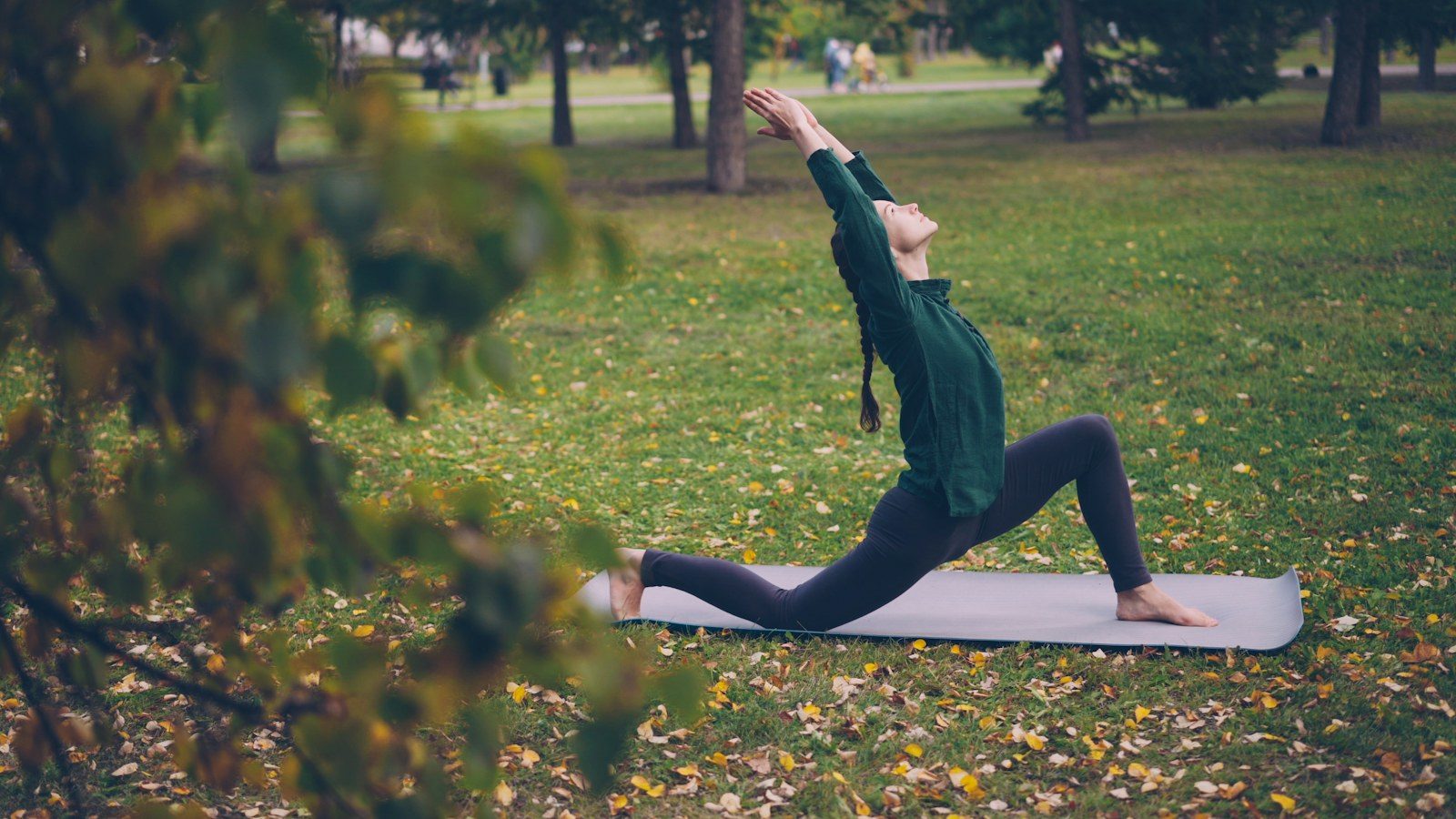 Mulher praticando yoga sozinha sobre um tapete num parque, alongando o corpo para cima